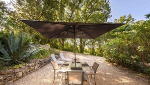 An outdoor patio with a table and chairs under a large umbrella, surrounded by gravel and greenery.