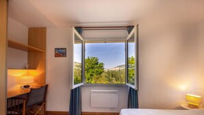 A bedroom with a large window, a desk with a chair, a bed, and a radiator.