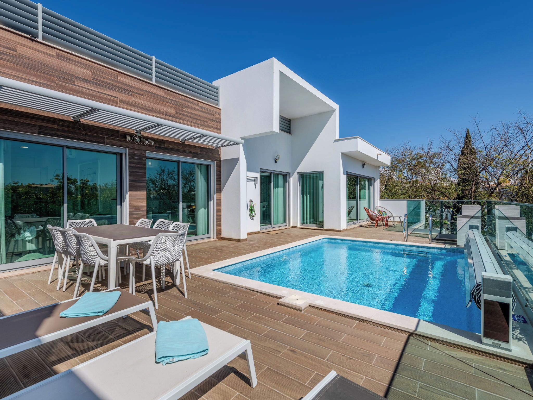 A modern outdoor pool area with a wooden deck, a dining set, and a lounge area.