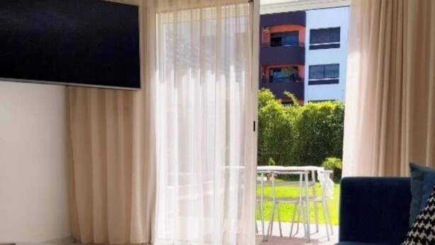 A modern living room with a blue sofa, a flat-screen TV mounted on the wall, and a sliding glass door leading to an outdoor area with a table and chairs. - Luxury Villa Hay Ryad (Rabat)