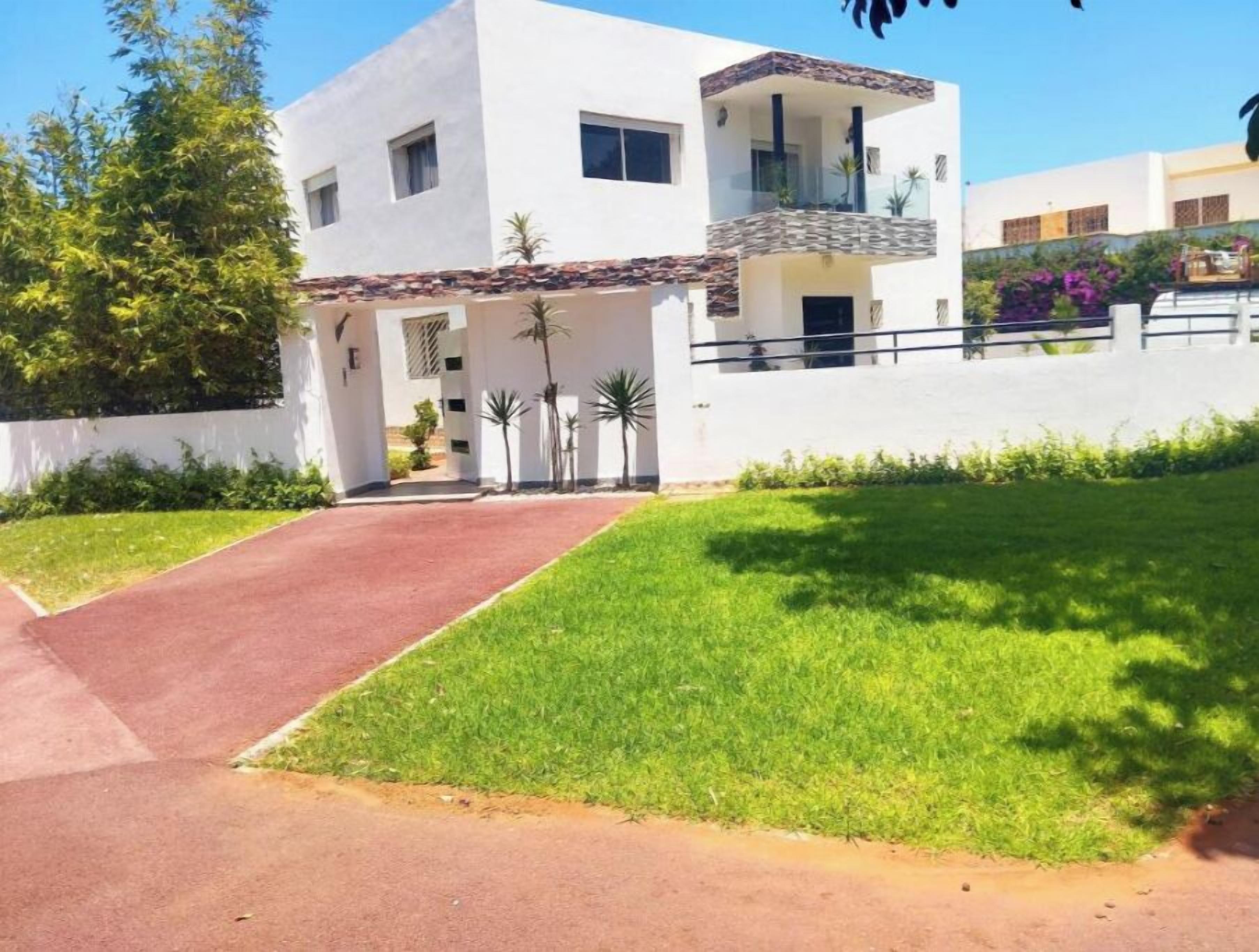 Une maison moderne à deux étages, dotée d’une allée pavée menant à la porte d’entrée, et entourée d’une pelouse bien entretenue et de petits palmiers.