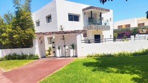 A modern two-story house with a paved driveway leading to a front door, surrounded by a well-maintained lawn and small palm trees. - Luxury Villa Hay Ryad (Rabat)