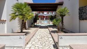 A narrow pathway leading to a building with a red facade and balconies. There are palm trees on either side of the path. - Luce del Mare by Interhome (Valledoria)