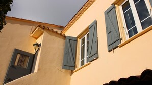 A building with two windows, each with wooden shutters, a door with a decorative metal grille, and a small balcony with a railing. - Romarin - EZE - Gîte (EZE)