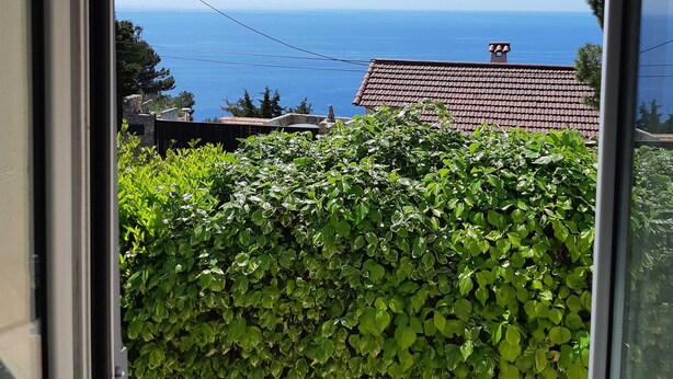 A view through a window of a building with a red-tiled roof, a green hedge, and a clear blue sky. - Romarin - EZE - Gîte (EZE)
