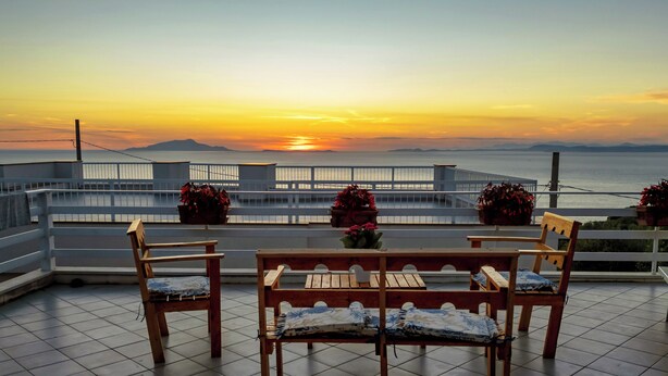 Una terraza con mesa y sillas de madera, plantas en macetas y vista al atardecer sobre el mar.