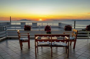 Una terraza con mesa y sillas de madera, plantas en macetas y vista al atardecer sobre el mar.
