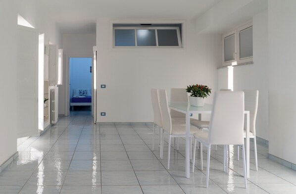 Un comedor moderno y bien iluminado con una mesa de vidrio, sillas blancas, una pequeña planta en maceta y vistas a otra habitación.