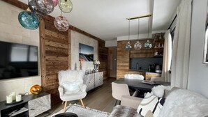 Una sala de estar moderna con televisor, sillas blancas, una mesa de comedor y una zona de cocina con armarios de madera y luces colgantes.