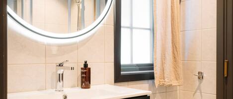 A bathroom with a round mirror, a sink, a bottle of shampoo, and a towel hanging by the door.