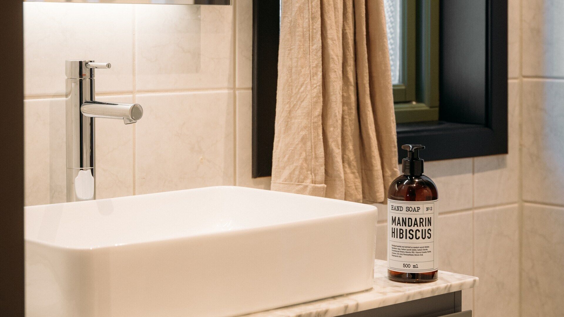 Une salle de bain avec un lavabo blanc, un flacon de savon liquide étiqueté « MANDARIN HIBISCUS », et une fenêtre à cadre noir.