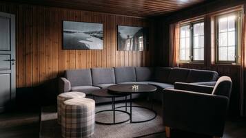 Ein gemütliches Zimmer mit einer Holzvertäfelung, einem grauen Ecksofa, einem kleinen runden Tisch und karierten Hocker.