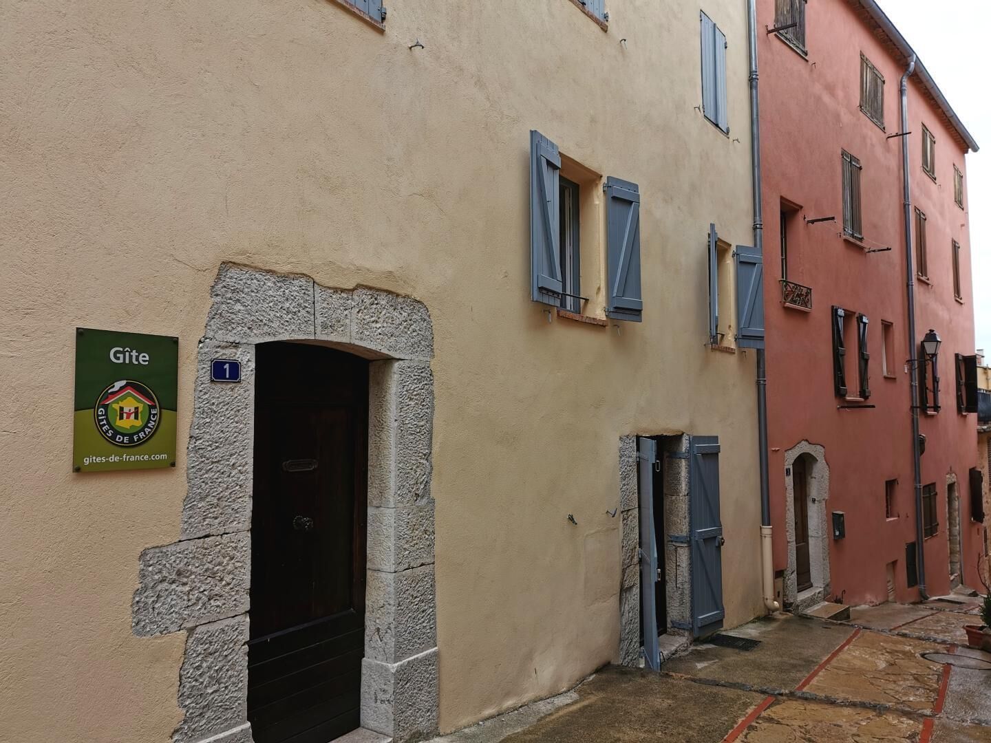 Eine enge Straße mit Gebäuden mit Stein- und Stuckfassaden, einem Schild mit der Aufschrift 'Gîte' und einer kleinen Tür.