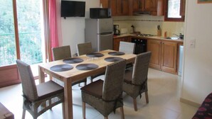 Een eetgedeelte met een houten tafel en rieten stoelen, een keuken met houten kasten, een koelkast en een televisie aan de muur.