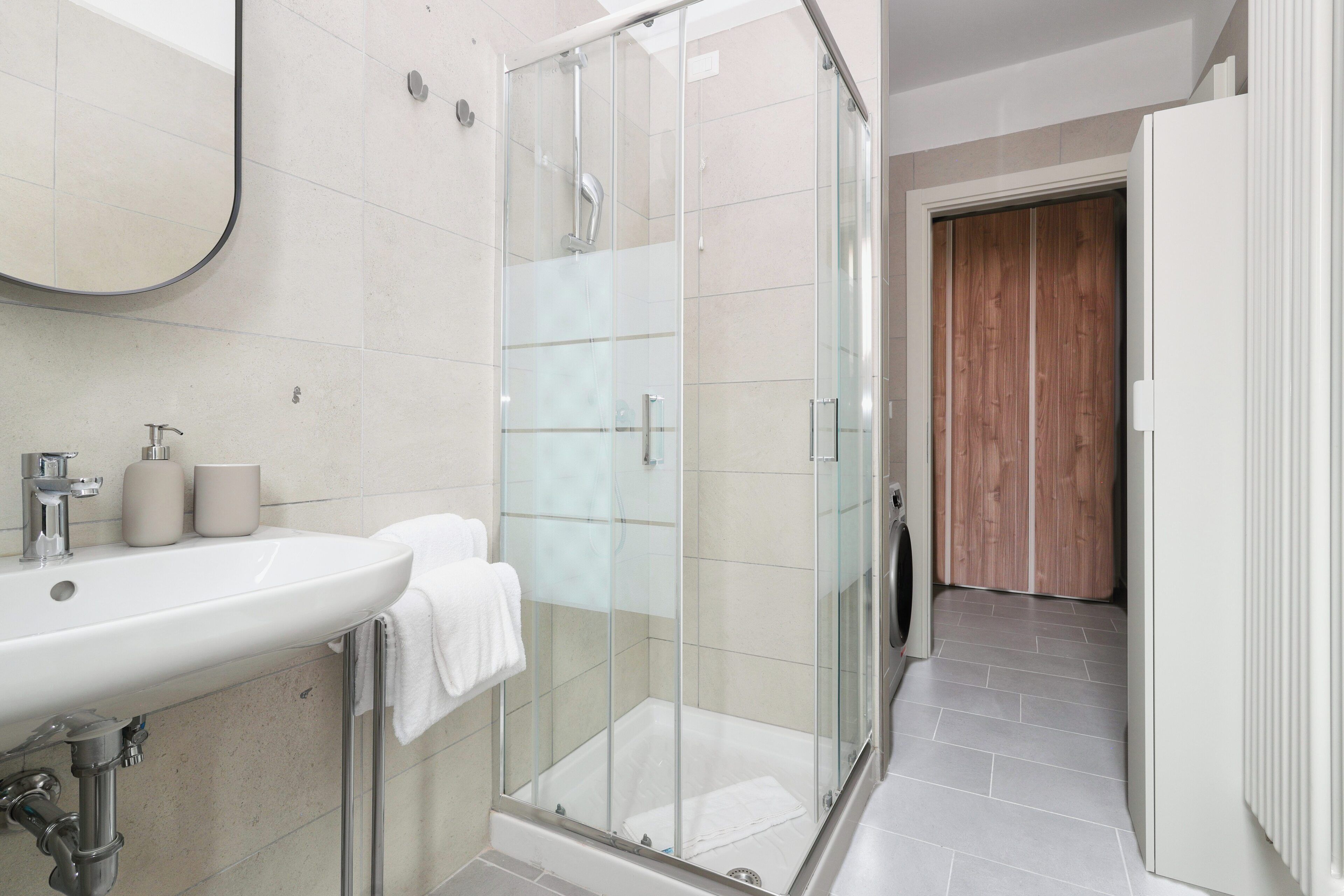 Un baño moderno con ducha de vidrio, un lavamanos blanco con espejo y una puerta de madera al fondo.