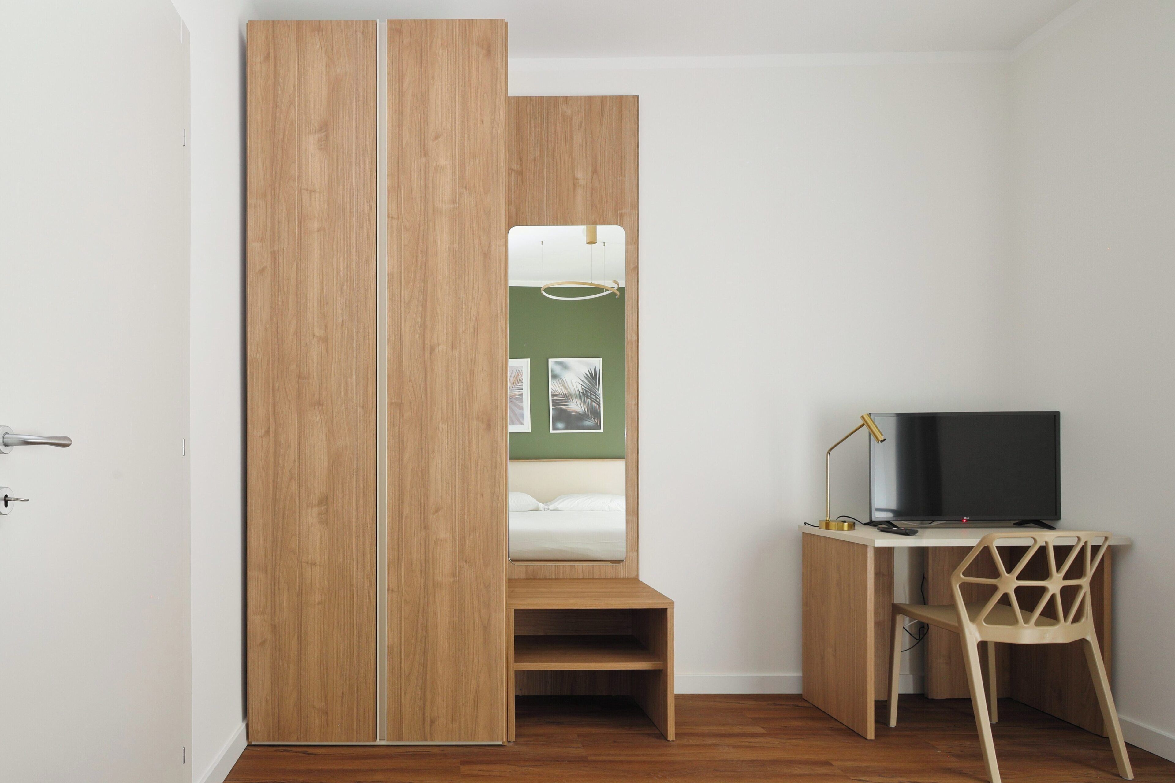 A room with a wooden wardrobe, a bed with a green wall, a desk with a chair, and a television.
