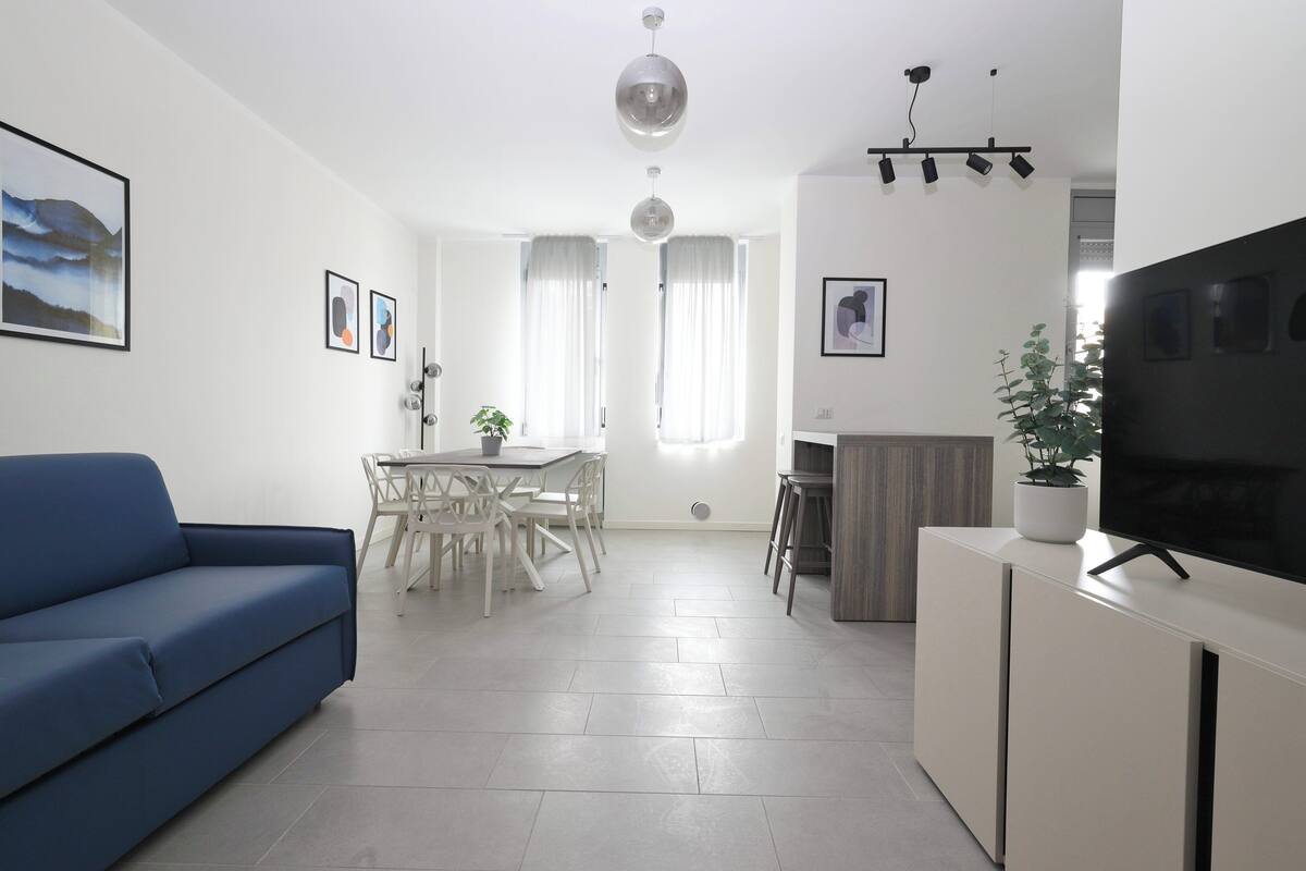 A modern living room with a blue sofa, a dining area with a table and chairs, a TV on a white cabinet, and artwork on the walls.