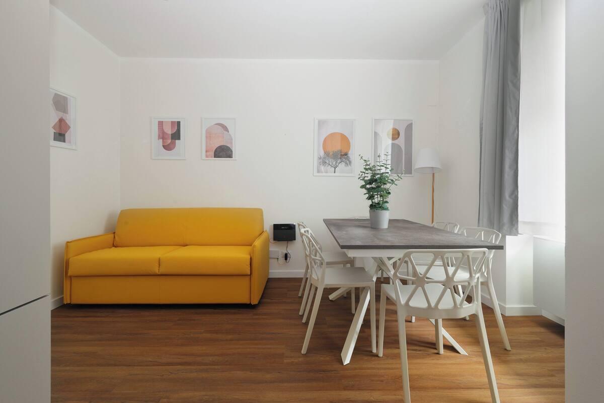 A modern living room with a yellow sofa, a dining table with chairs, and a potted plant.