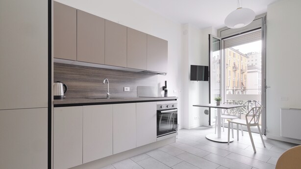 Una cocina moderna con armarios blancos, una encimera negra y un horno empotrado. Hay una zona de comedor con una mesa y sillas, y un ventanal con vistas a edificios.