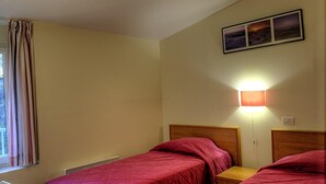 A room with two single beds, a window with curtains, a wall-mounted light, and a picture above the beds.