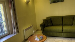 A living room with a green sofa, a table with books and a glass, a radiator, a window with curtains, and a wall-mounted light fixture.
