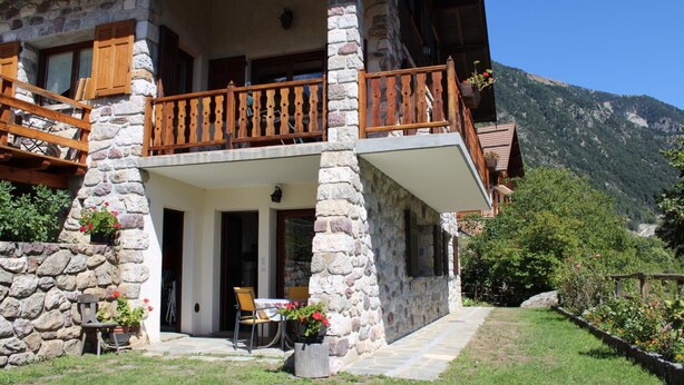 Un edificio de piedra con balcón, una mesa y sillas, y montañas al fondo.