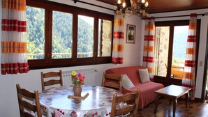 Un comedor acogedor con una mesa redonda, sillas y un sofá. Hay ventanas amplias con cortinas, una araña y vistas a un área boscosa.