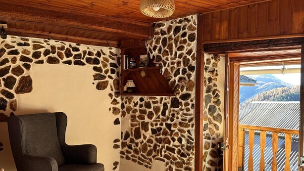 Une pièce avec un mur en pierre, un plafond en bois, une chaise grise et une porte en bois donnant sur un balcon avec vue sur les montagnes.