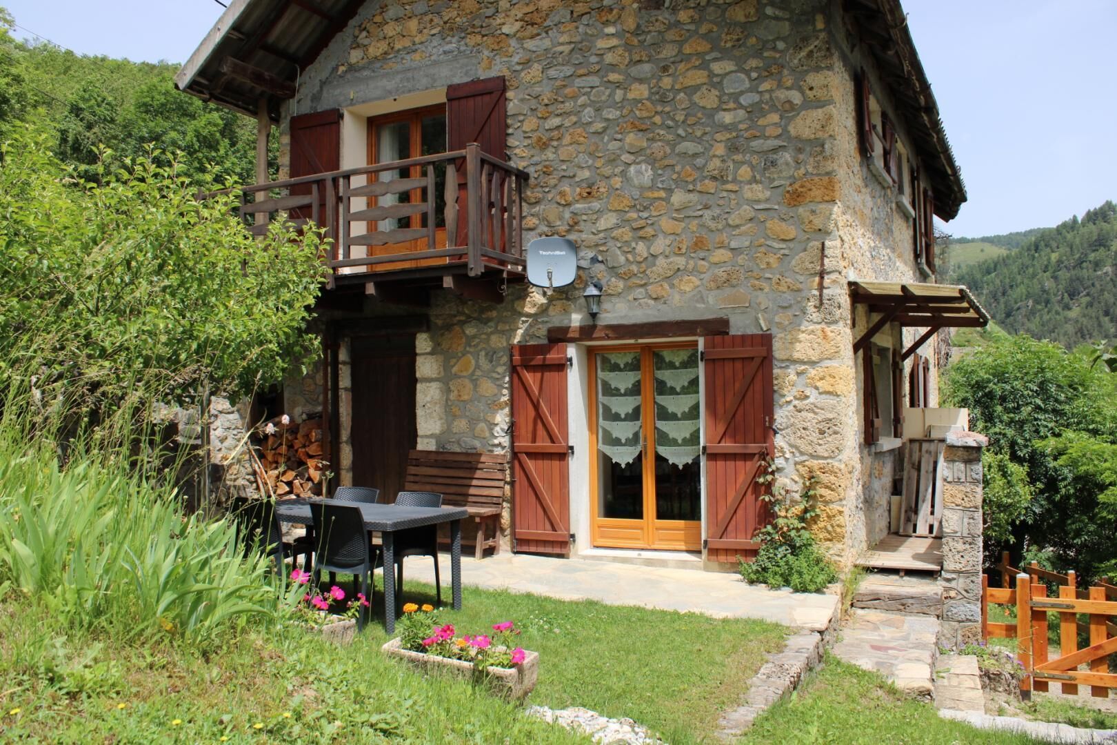A stone house with a balcony, a wooden door, and a small garden with flowers.