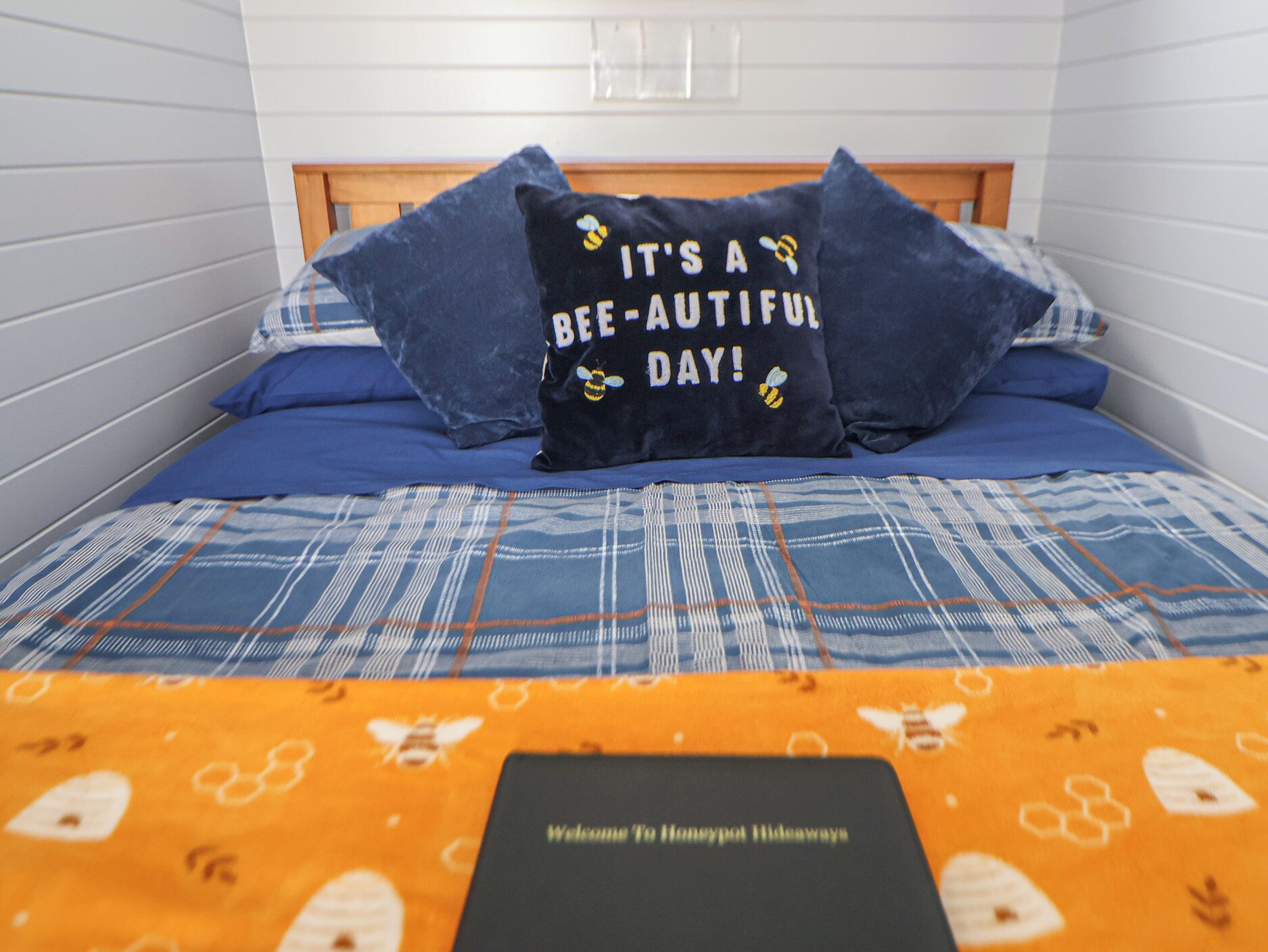 Ein Bett mit einer blau-weißen Karodecke, zwei Kissen mit Bienenmuster und einem Willkommensschild darauf.
