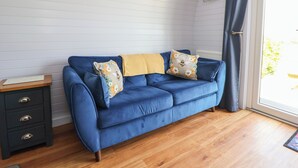 A blue sofa with floral pillows, a wooden side table, and a view of greenery through a window. - Honeypot Hideaways - Manuka (Chester)