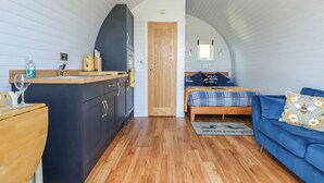 A compact room with a kitchenette, a bed with a blue and yellow plaid bedspread, and a blue sofa with a floral pillow. - Honeypot Hideaways - Manuka (Chester)