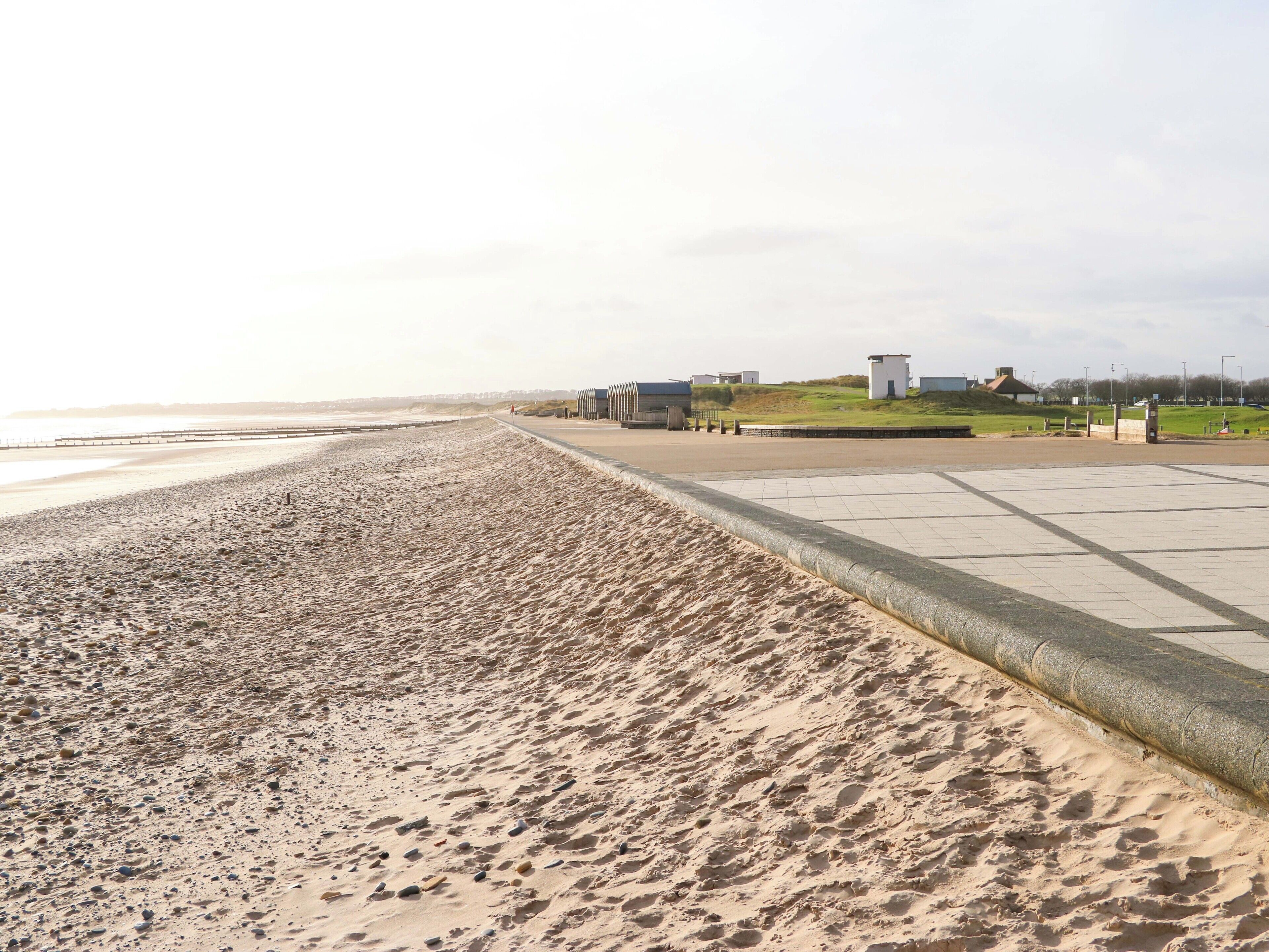 A beach with a concrete walkway, a grassy area, and some buildings in the background.