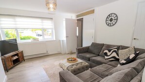 Ein Wohnzimmer mit grauem Sofa, einem Fernseher auf einem Holzboden und einem großen Fenster mit Jalousien.