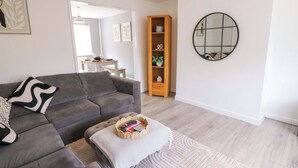 Ein modernes Wohnzimmer mit einem grauen Ecksofa, einem kleinen Hocker mit Korb, einem Holzregal und einem runden Spiegel an der Wand.