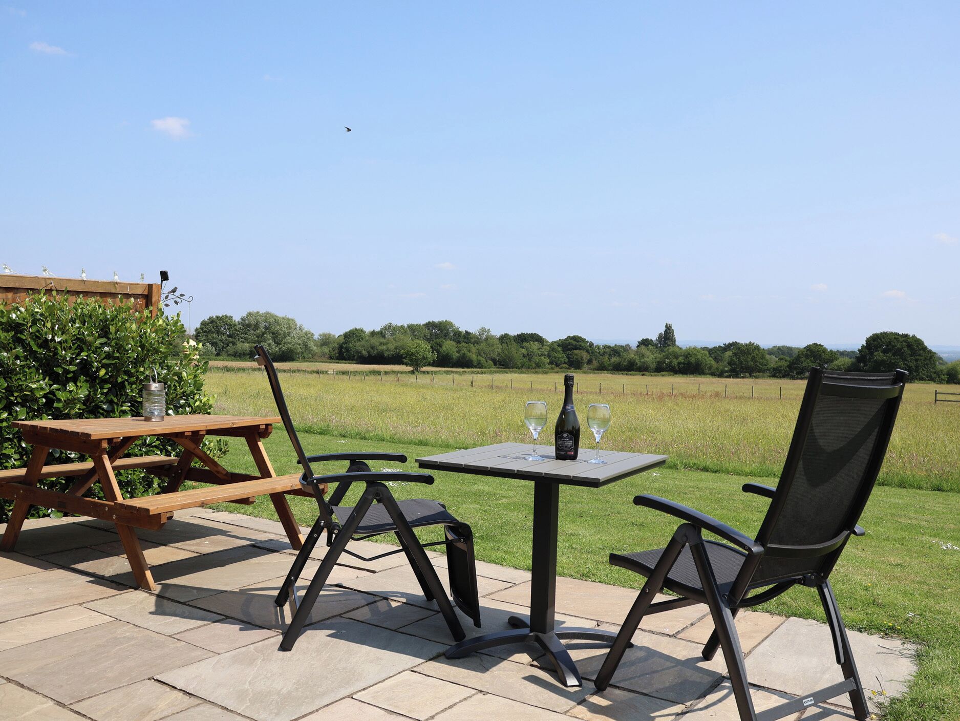 An outdoor seating area with a picnic table and chairs, a small table with two wine glasses and a bottle, and a view of a grassy field and trees.