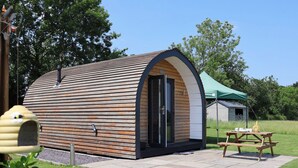 A small, wooden, semi-circular structure with a door and windows, situated on a paved patio with a picnic table nearby. - Honeypot Hideaways - Clover (Chester)