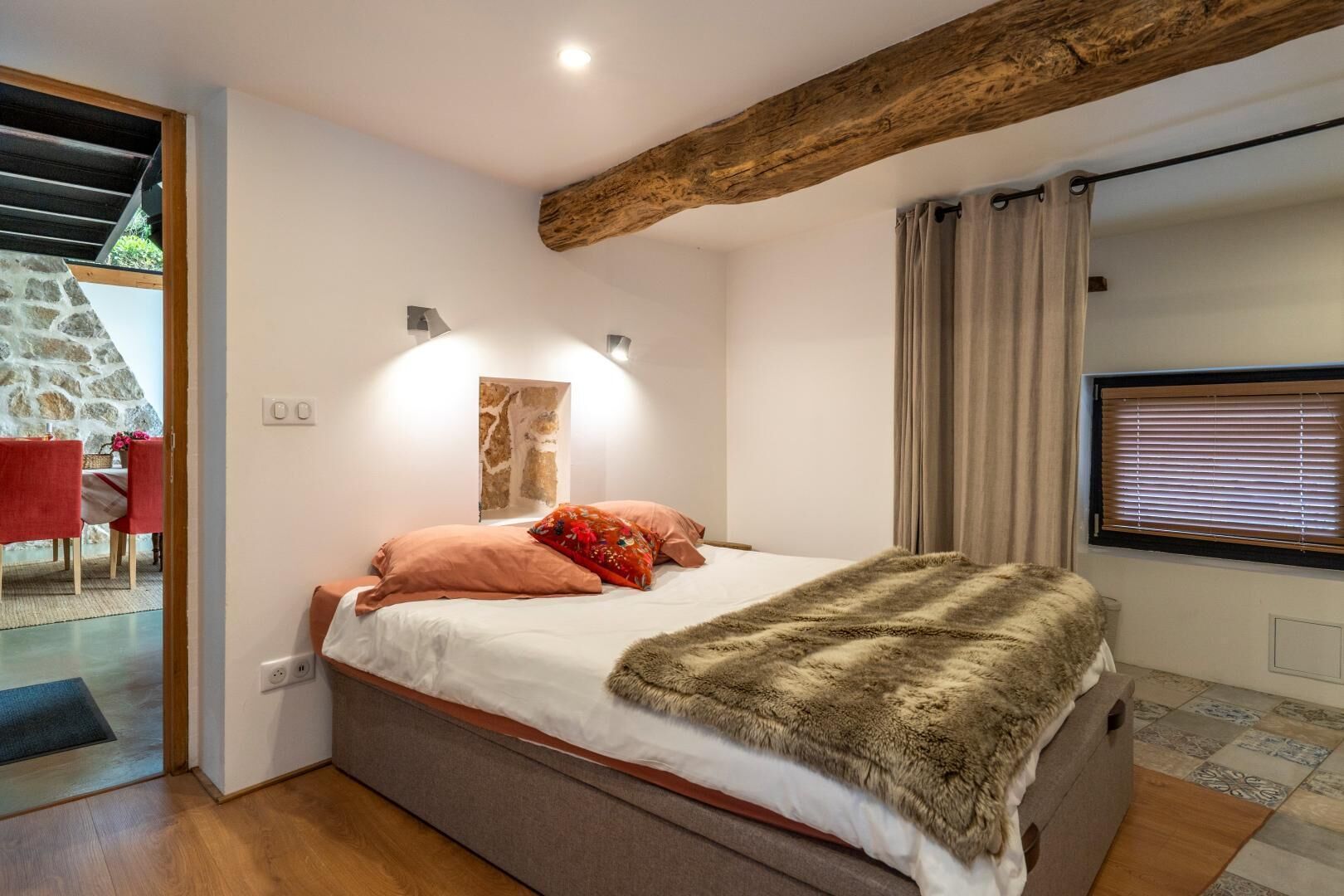 A bedroom with a bed, a stone wall, a wooden ceiling beam, a window with curtains, and a red chair in the adjacent room.