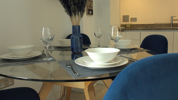 Una mesa de comedor dispuesta con cuencos blancos, copas de vino y cubiertos, con un jarrón de plantas secas sobre la mesa.