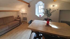 A cozy room with a wooden ceiling, a dining table with chairs, a sofa, a floor lamp, and a window with a view.