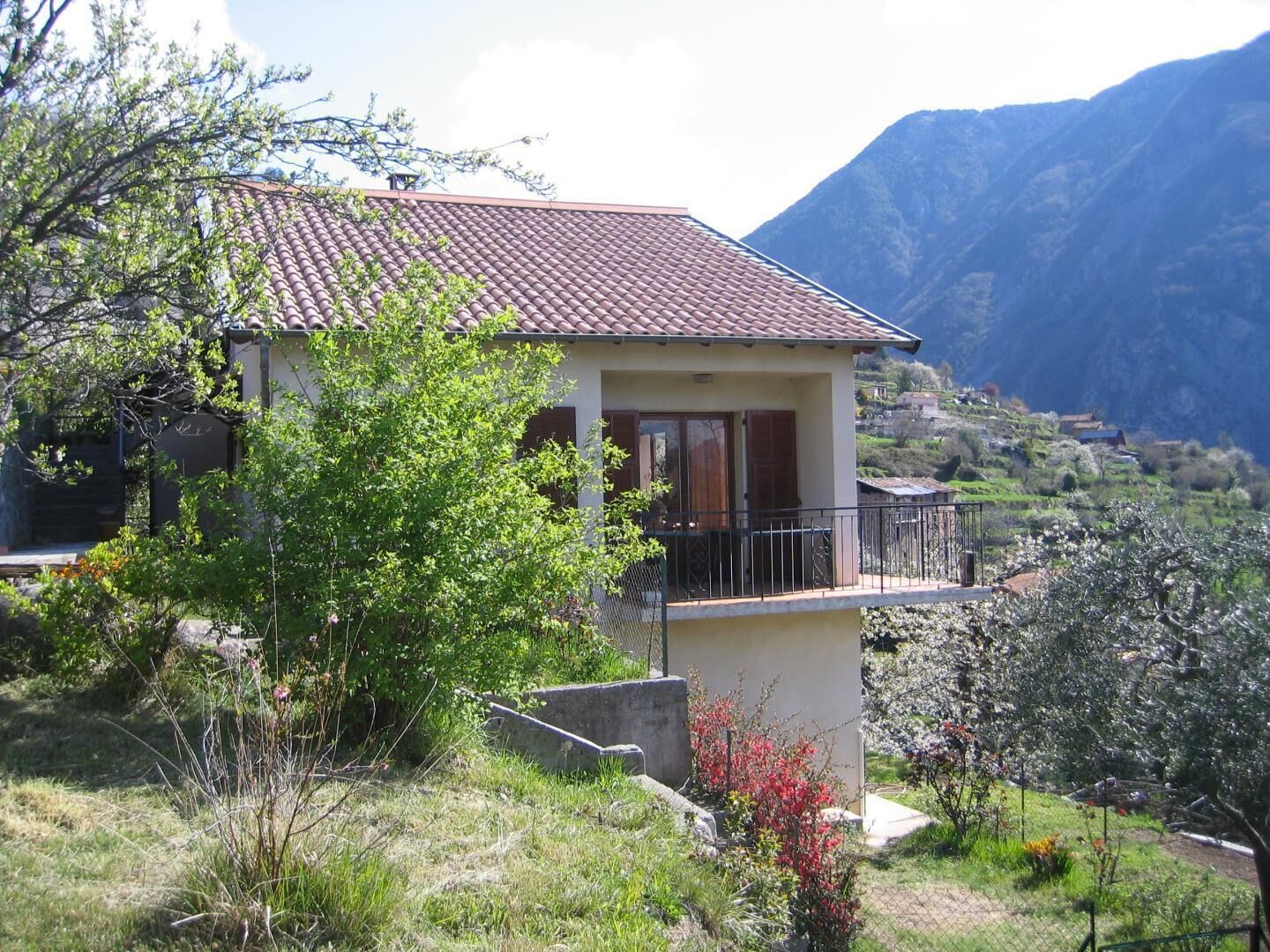 Une maison à deux étages avec un balcon, entourée de verdure et avec des montagnes en arrière-plan.