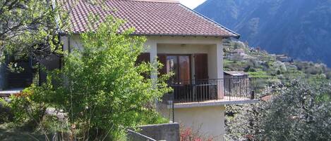 Une maison à deux étages avec un balcon, entourée de verdure et avec des montagnes en arrière-plan.