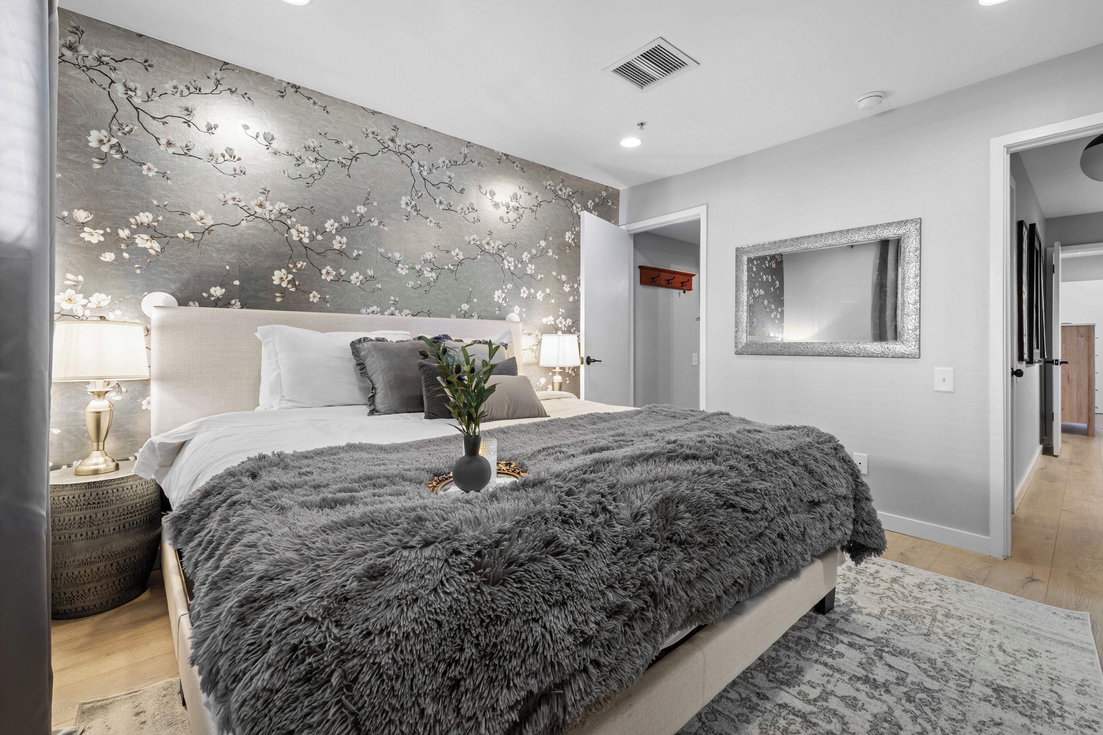 A bedroom with a bed, a nightstand, a lamp, a mirror, and a wall with a floral pattern.