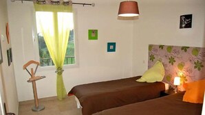 Een slaapkamer met twee eenpersoonsbedden, een raam met dunne gordijnen, een bedlampje en een klein tafeltje.