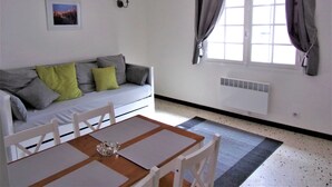 Une pièce avec un canapé blanc, une table à manger avec des chaises, une fenêtre avec des rideaux, un radiateur fixé au mur et un tableau accroché au mur.