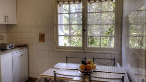 Une cuisine avec des meubles blancs, une table à manger avec des chaises, un panier de fruits et une fenêtre donnant sur un paysage verdoyant.