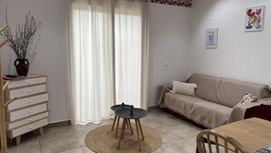 Una sala de estar con un sofá, una mesa de comedor, una mesa auxiliar, un jarrón con ramas y una cortina.