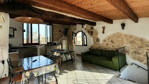 A rustic interior with a stone wall, wooden ceiling beams, a dining table with chairs, a green sofa, and a kitchen area with a window. - La Maison du Chateau (SAINT-JEANNET)