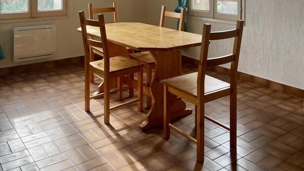 A dining area with a wooden table and chairs, a television mounted on the wall, and windows with curtains.
