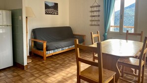 A cozy room with a wooden floor, a grey sofa, a wooden dining table with chairs, a window with curtains, and a wall-mounted decoration.
